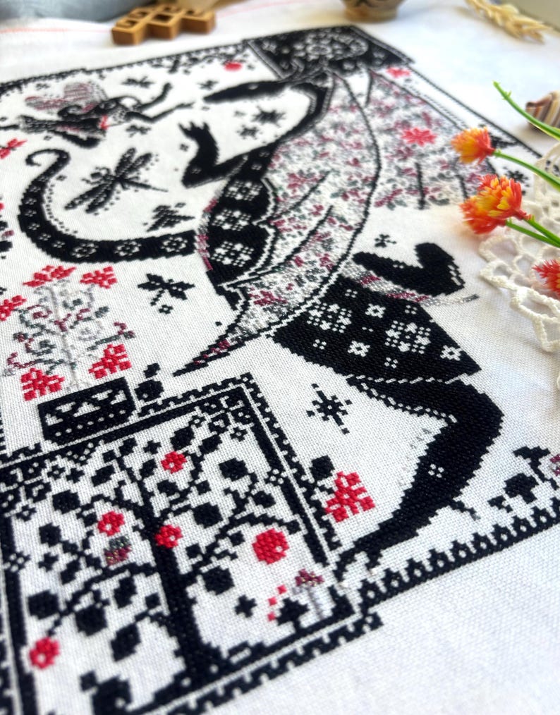 May include: A white fabric piece featuring a detailed cross-stitch embroidery pattern in black and red thread. The design depicts a large winged creature, a dragonfly, and floral motifs arranged within a decorative border on the textile.