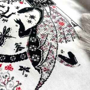 May include: A cross-stitch embroidery on white fabric featuring a black dragon with intricate patterns. The dragon has wings filled with small red and gray geometric motifs. Surrounding the dragon are black star-like shapes, dragonfly silhouettes, and decorative borders.