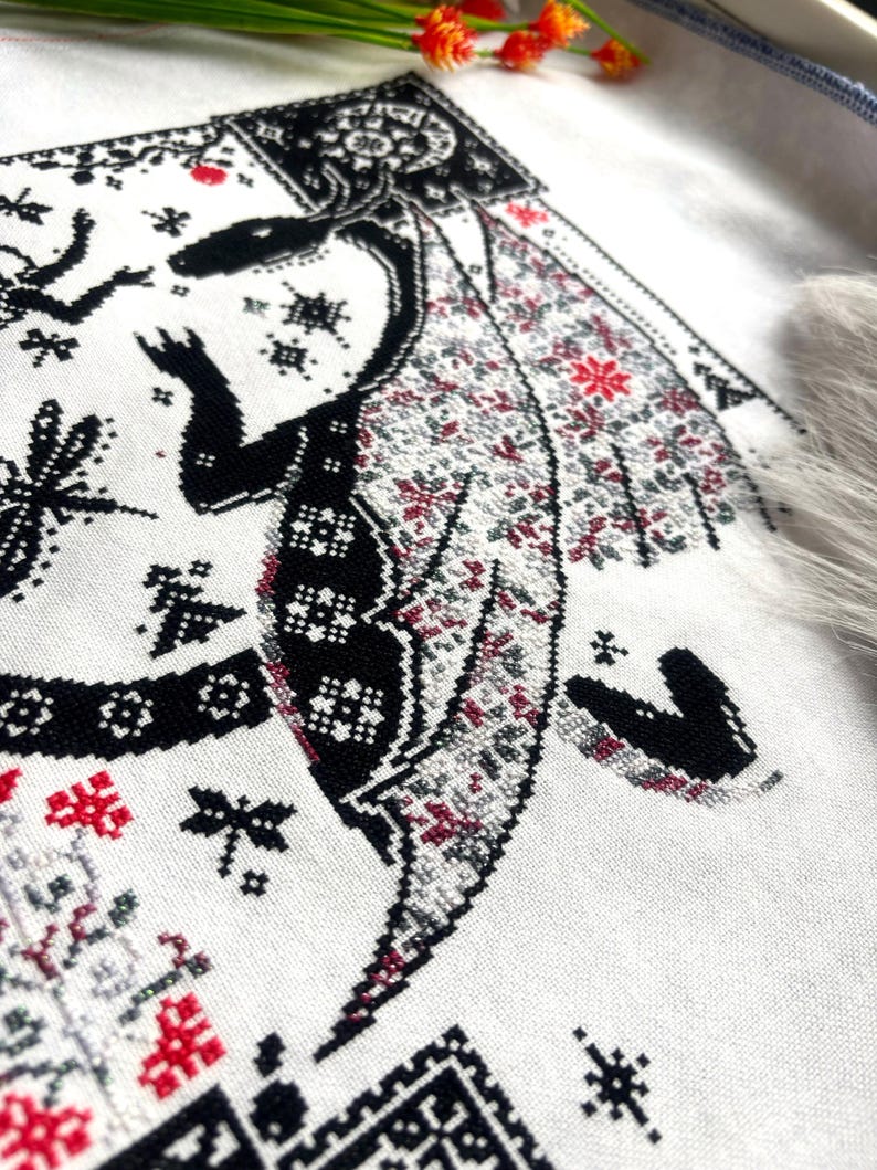 May include: A cross-stitch embroidery on white fabric featuring a black dragon with intricate patterns. The dragon has wings filled with small red and gray geometric motifs. Surrounding the dragon are black star-like shapes, dragonfly silhouettes, and decorative borders.