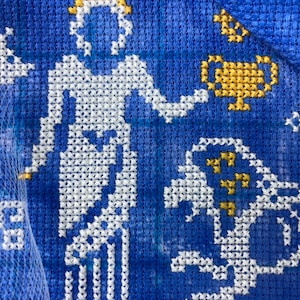 May include: Close-up of a blue cross-stitch fabric featuring a white figure holding a gold cup. The figure has a gold crown. Additional gold and white cross-stitch details include stars and a horse-like creature.