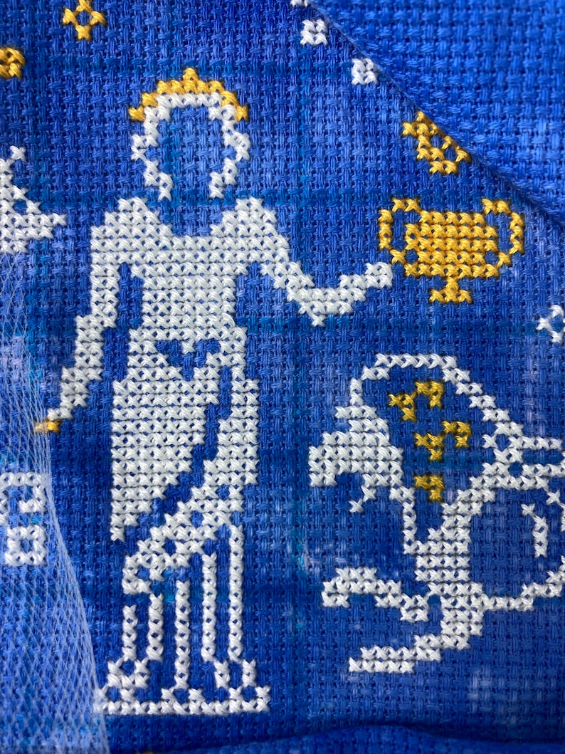 May include: Close-up of a blue cross-stitch fabric featuring a white figure holding a gold cup. The figure has a gold crown. Additional gold and white cross-stitch details include stars and a horse-like creature.