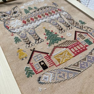 May include: Cross-stitch art of a cat with a patterned design, set against a winter village scene. The cat features a detailed pattern in shades of brown, red, and white. The village includes houses, trees, and a bell, all framed in a wooden border.