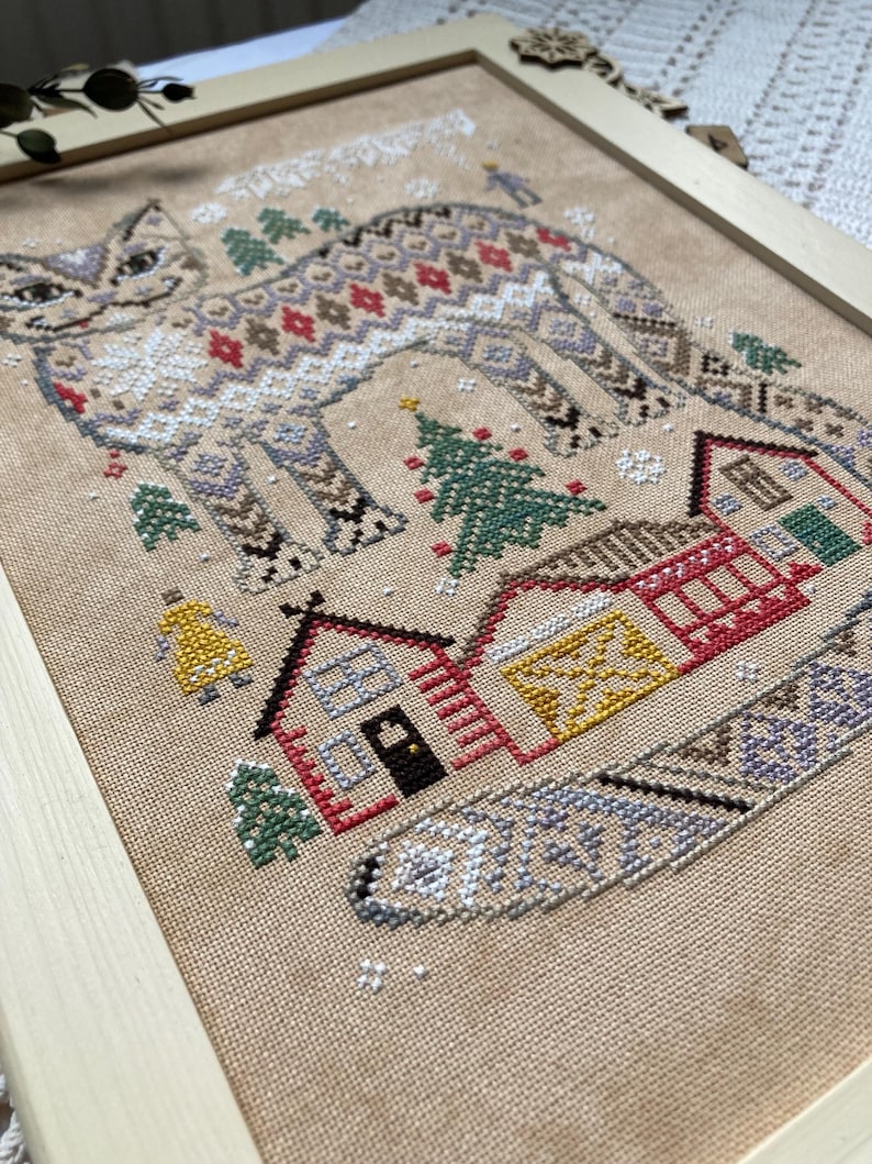 May include: Cross-stitch art of a cat with a patterned design, set against a winter village scene. The cat features a detailed pattern in shades of brown, red, and white. The village includes houses, trees, and a bell, all framed in a wooden border.
