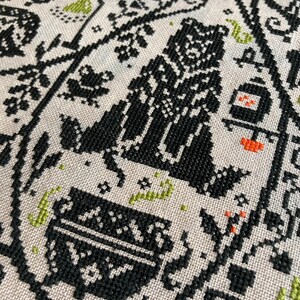 May include: Close-up of a beige fabric with a black cross-stitch pattern. The design includes stylized figures, plants, and geometric shapes, with accents of orange and green. The overall aesthetic is folk art.