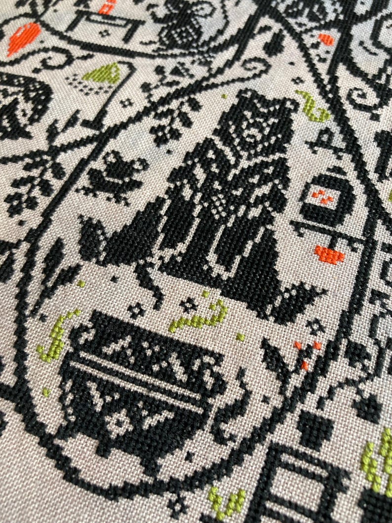 May include: Close-up of a beige fabric with a black cross-stitch pattern. The design includes stylized figures, plants, and geometric shapes, with accents of orange and green. The overall aesthetic is folk art.