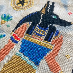 Puede incluir: Punto de cruz de Anubis, el dios egipcio del m&aacute;s all&aacute;, en hilo negro, azul, dorado y naranja. La obra presenta una representaci&oacute;n detallada de Anubis con cabeza de chacal, patrones intrincados y un dise&ntilde;o celestial en el fondo.