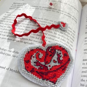 Peut inclure: Marque-page en forme de c&oelig;ur avec un motif de dragon rouge brod&eacute; sur fond gris. Il est orn&eacute; d'un ruban rouge. Une petite bouteille en verre avec un bouchon en li&egrave;ge est en arri&egrave;re-plan.