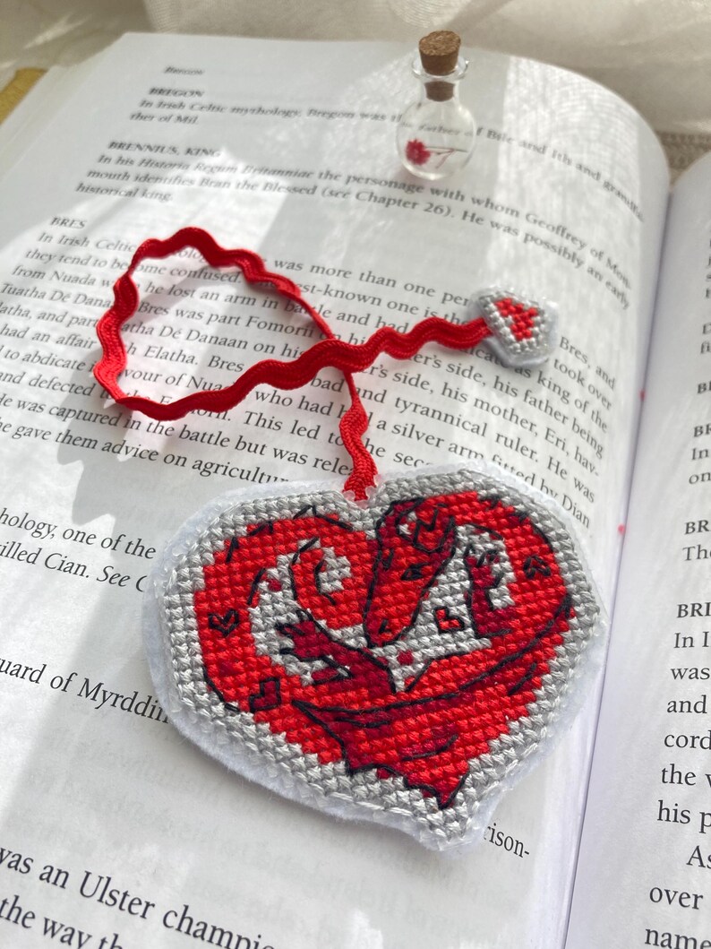Peut inclure: Marque-page en forme de c&oelig;ur avec un motif de dragon rouge brod&eacute; sur fond gris. Il est orn&eacute; d'un ruban rouge. Une petite bouteille en verre avec un bouchon en li&egrave;ge est en arri&egrave;re-plan.