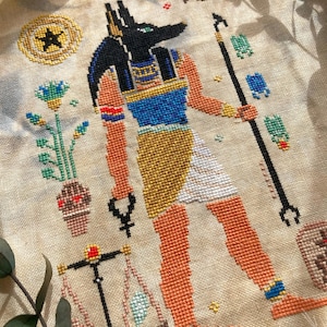 Puede incluir: Punto de cruz del dios egipcio Anubis, representado con cabeza de chacal negra, piel naranja y detalles dorados y azules. La obra incluye balanzas, un b&aacute;culo y otros s&iacute;mbolos egipcios, bordados sobre tela beige.