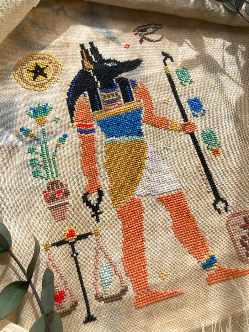 Puede incluir: Punto de cruz del dios egipcio Anubis, representado con cabeza de chacal negra, piel naranja y detalles dorados y azules. La obra incluye balanzas, un b&aacute;culo y otros s&iacute;mbolos egipcios, bordados sobre tela beige.