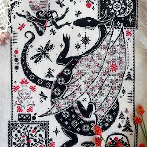 May include: A cross-stitch embroidery on white fabric featuring a large black dragon with patterned wings, a small fairy, dragonflies, and a moon. The design includes red and black floral motifs, trees, and geometric borders in a rectangular layout.