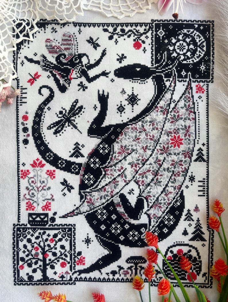 May include: A cross-stitch embroidery on white fabric featuring a large black dragon with patterned wings, a small fairy, dragonflies, and a moon. The design includes red and black floral motifs, trees, and geometric borders in a rectangular layout.