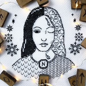May include: A black and white cross-stitch embroidery of a woman's face with a necklace featuring the letter N. The embroidery is surrounded by a string of lights and wooden runes.