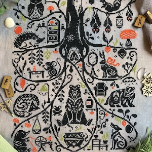 May include: A detailed cross-stitch design featuring a central tree with various woodland creatures and objects. The design includes an owl, squirrel, bats, bear, and other animals, with orange and green accents on a neutral background.