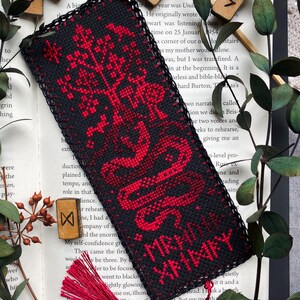 K&ouml;nnte beinhalten: Schwarz-roter, bestickter Lesezeichen mit Quaste. Das Lesezeichen zeigt ein rotes Baum- und Schlangenmuster mit dem Text "MR. XANNAY" in Rot. Das Lesezeichen liegt auf einem offenen Buch mit Holzrunen.