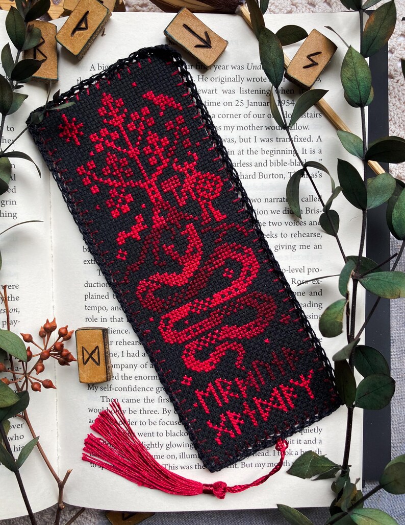 K&ouml;nnte beinhalten: Schwarz-roter, bestickter Lesezeichen mit Quaste. Das Lesezeichen zeigt ein rotes Baum- und Schlangenmuster mit dem Text "MR. XANNAY" in Rot. Das Lesezeichen liegt auf einem offenen Buch mit Holzrunen.