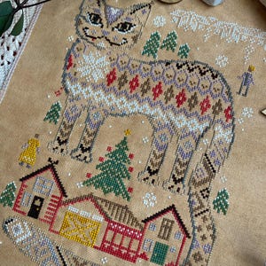 May include: Cross-stitch design featuring a stylized cat with a patterned body, surrounded by small houses, trees, and snowflakes. The color palette includes beige, brown, red, green, and white, creating a cozy, wintery scene. The design is on a tan fabric.