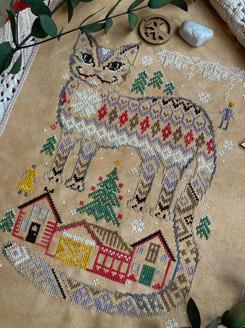 May include: Cross-stitch design featuring a stylized cat with a patterned body, surrounded by small houses, trees, and snowflakes. The color palette includes beige, brown, red, green, and white, creating a cozy, wintery scene. The design is on a tan fabric.