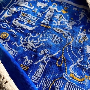 May include: Close-up of a blue textile with white and gold cross-stitch embroidery. The embroidery depicts figures, boats, and geometric patterns. The textile is framed by a white crocheted border.