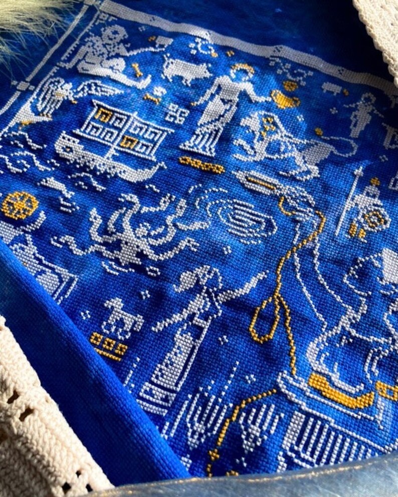 May include: Close-up of a blue textile with white and gold cross-stitch embroidery. The embroidery depicts figures, boats, and geometric patterns. The textile is framed by a white crocheted border.