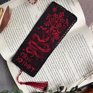 K&ouml;nnte beinhalten: Schwarz-rotes besticktes Lesezeichen mit Quaste. Das Lesezeichen zeigt ein detailliertes Design eines Baumes und einer Schlange mit dem Text "XRHARY XRHMFY". Das Lesezeichen liegt auf einem offenen Buch.
