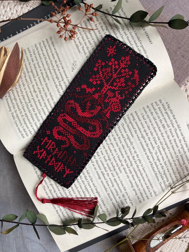 K&ouml;nnte beinhalten: Schwarz-rotes besticktes Lesezeichen mit Quaste. Das Lesezeichen zeigt ein detailliertes Design eines Baumes und einer Schlange mit dem Text "XRHARY XRHMFY". Das Lesezeichen liegt auf einem offenen Buch.