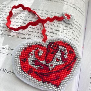 Peut inclure: Marque-page fait main en forme de c&oelig;ur avec un motif de dragon rouge. Le marque-page est en tissu point de croix avec un ruban rouge. Le dragon est rouge avec des d&eacute;tails gris, et le bord est gris.
