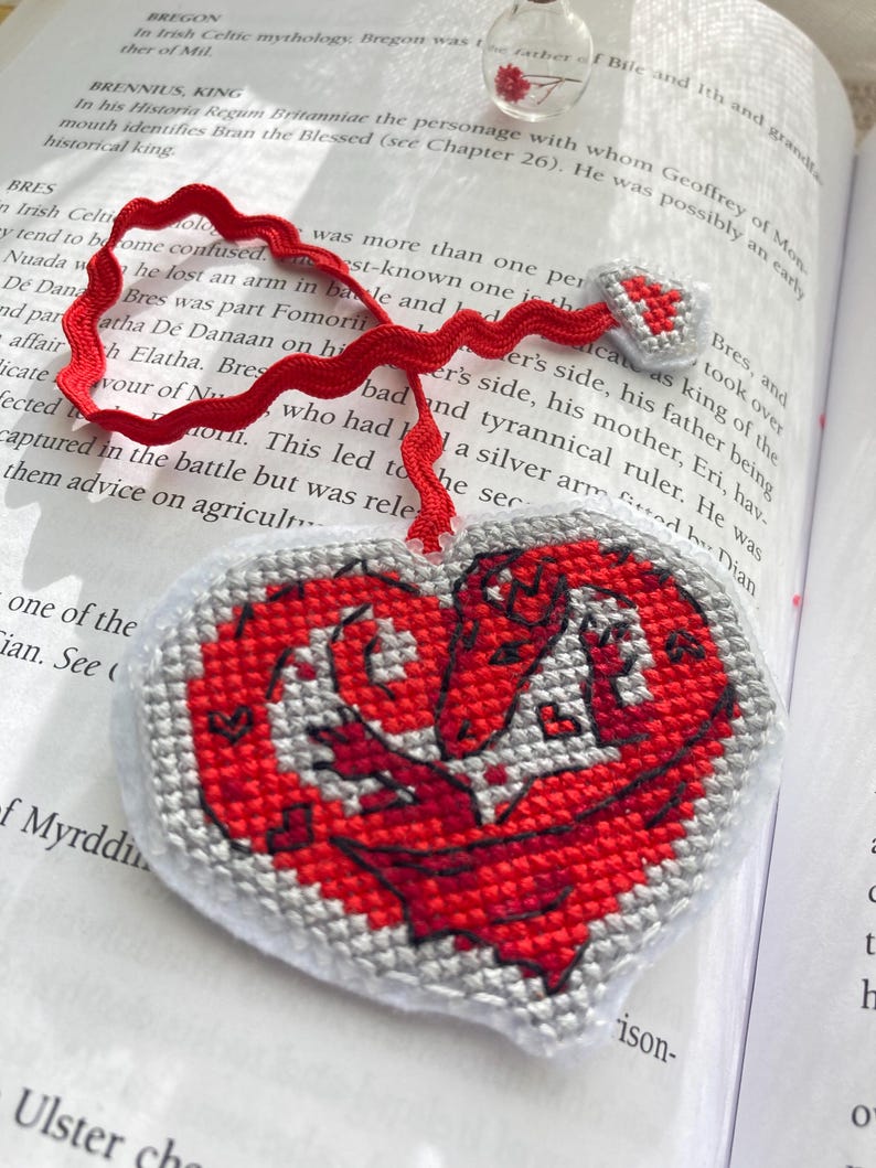 Peut inclure: Marque-page fait main en forme de c&oelig;ur avec un motif de dragon rouge. Le marque-page est en tissu point de croix avec un ruban rouge. Le dragon est rouge avec des d&eacute;tails gris, et le bord est gris.