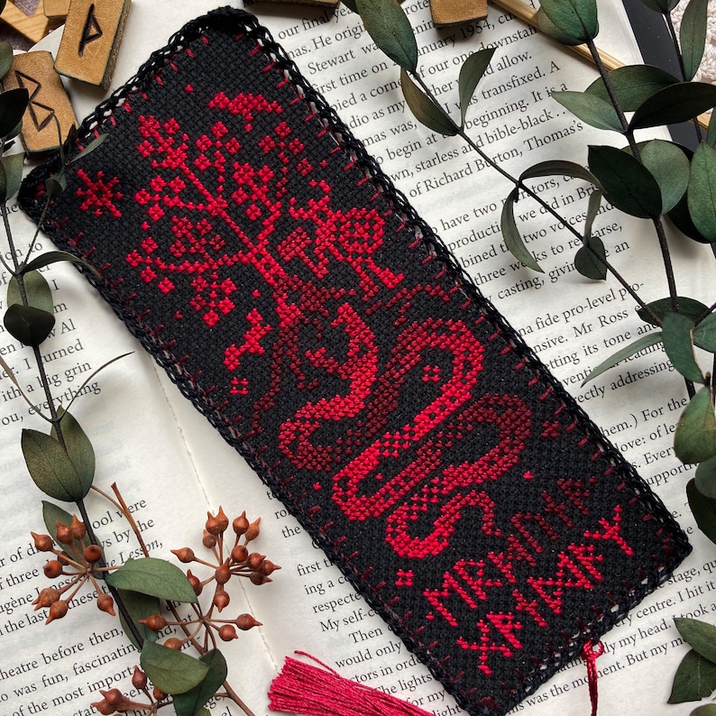 K&ouml;nnte beinhalten: Ein schwarz-rotes Kreuzstich-Lesezeichen mit einem Baum- und Schlangenmuster. Das Lesezeichen tr&auml;gt den Text "MRH XHARY" in Rot. Das Lesezeichen ist rechteckig mit einem schwarzen Rand und einer roten Quaste.