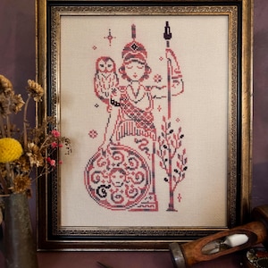 May include: Framed cross-stitch artwork featuring a figure holding a spear and an owl, in shades of red, black, and cream. The artwork is in a gold-toned frame. The image includes a vase of dried flowers and a pair of scissors.