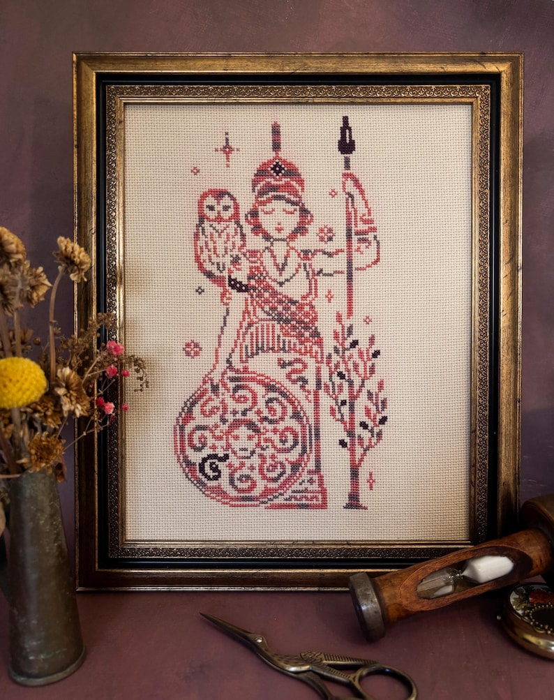 May include: Framed cross-stitch artwork featuring a figure holding a spear and an owl, in shades of red, black, and cream. The artwork is in a gold-toned frame. The image includes a vase of dried flowers and a pair of scissors.
