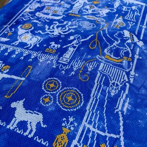 May include: A detailed cross-stitch pattern on blue fabric. The design features white and gold elements, including figures, architectural details, and geometric shapes. The overall aesthetic is reminiscent of ancient art and mythology.