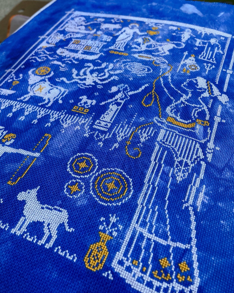 May include: A detailed cross-stitch pattern on blue fabric. The design features white and gold elements, including figures, architectural details, and geometric shapes. The overall aesthetic is reminiscent of ancient art and mythology.