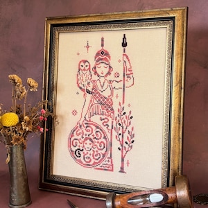May include: Framed cross-stitch artwork featuring a figure in red and black, holding a spear and owl. The artwork is in a gold-toned frame with a black inner border. The background is a neutral beige color.