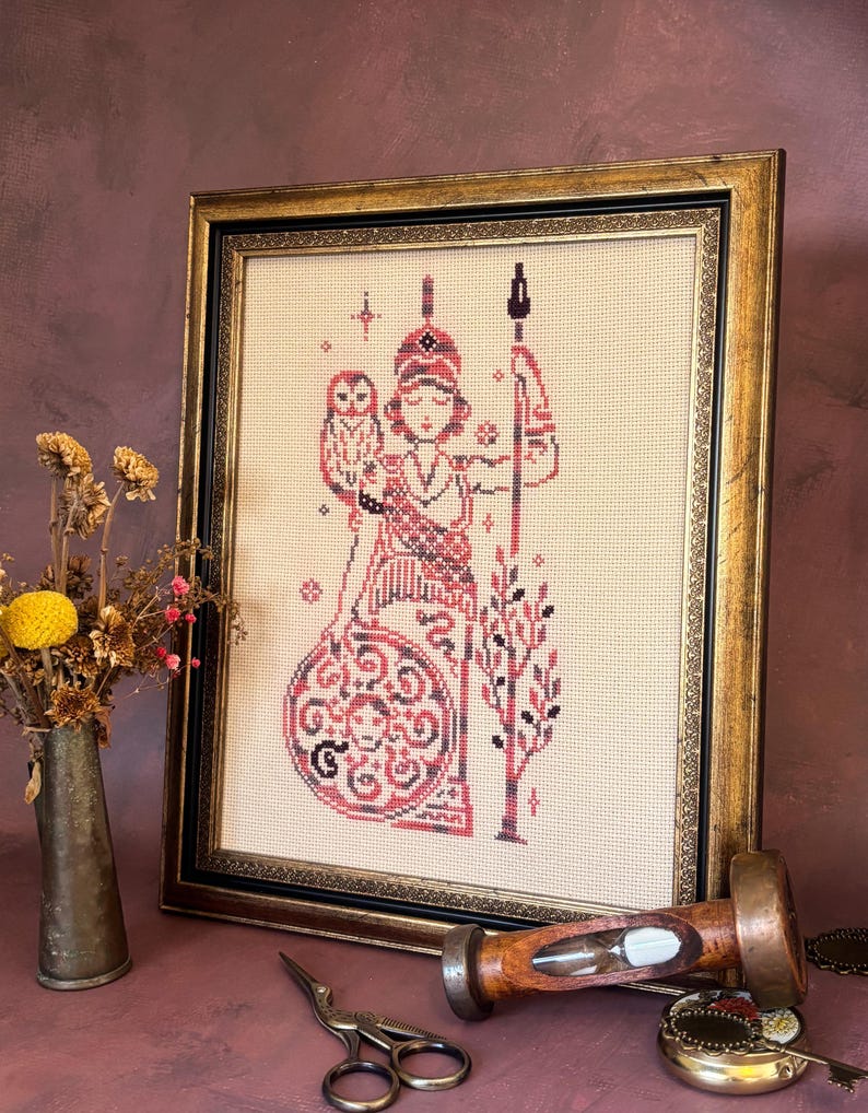 May include: Framed cross-stitch artwork featuring a figure in red and black, holding a spear and owl. The artwork is in a gold-toned frame with a black inner border. The background is a neutral beige color.