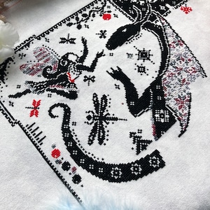 May include: A cross-stitch embroidery on white fabric featuring a black silhouette of a dragon and a small winged figure. The design includes intricate geometric patterns, small red accents, and a dragonfly motif, all rendered in black thread.