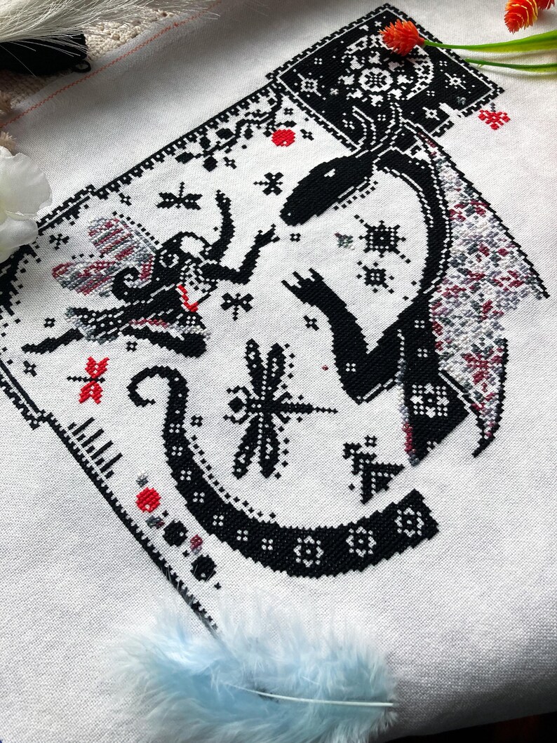 May include: A cross-stitch embroidery on white fabric featuring a black silhouette of a dragon and a small winged figure. The design includes intricate geometric patterns, small red accents, and a dragonfly motif, all rendered in black thread.