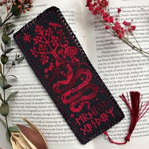 K&ouml;nnte beinhalten: Schwarz-roter bestickter Lesezeichen mit einem Baum- und Schlangenmuster. Das Lesezeichen hat einen schwarzen Rand und eine rote Quaste. Der Text "MRADI XHMXY" ist auf dem Lesezeichen zu sehen.