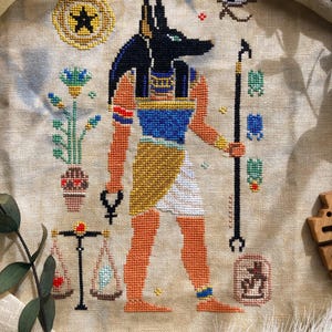 Puede incluir: Punto de cruz de Anubis, el dios con cabeza de chacal, en negro, azul, naranja y dorado. La obra de arte incluye un bast&oacute;n, balanzas y otros s&iacute;mbolos egipcios. El fondo es de tela beige claro.