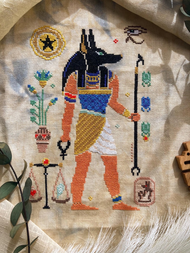 Puede incluir: Punto de cruz de Anubis, el dios con cabeza de chacal, en negro, azul, naranja y dorado. La obra de arte incluye un bast&oacute;n, balanzas y otros s&iacute;mbolos egipcios. El fondo es de tela beige claro.