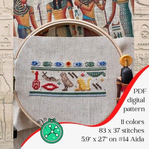 May include: A cross-stitch pattern featuring Egyptian hieroglyphs, displayed in a wooden embroidery hoop. The design includes symbols in red, blue, yellow, and green. The text "PDF digital pattern" is visible, along with stitch and size details: 5.9" x 2.7".