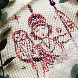 May include: Cross-stitch embroidery featuring a figure with an owl, rendered in shades of red, purple, and white on a cream-colored fabric. The design includes stars and a spear, with a rustic, handmade aesthetic.