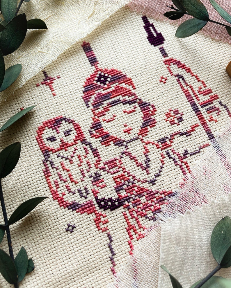 May include: Cross-stitch embroidery featuring a figure with an owl, rendered in shades of red, purple, and white on a cream-colored fabric. The design includes stars and a spear, with a rustic, handmade aesthetic.