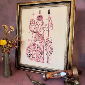 May include: Framed cross-stitch artwork featuring a figure in red and black, holding a spear and shield, with an owl. The artwork is in a gold-colored frame. Other items include a small vase with dried flowers, scissors, and a wooden hourglass.