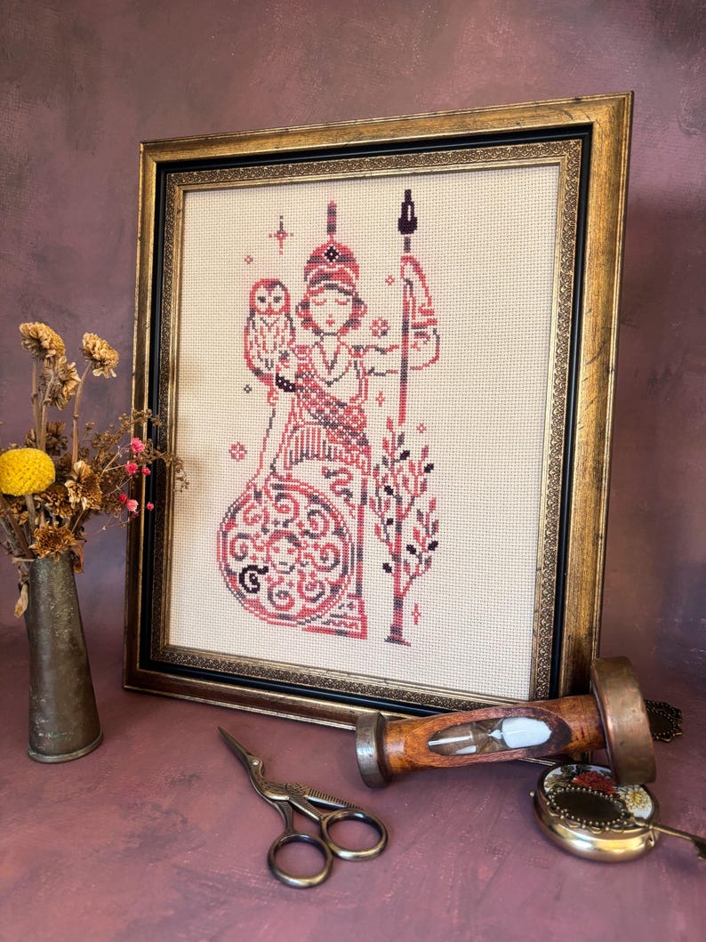 May include: Framed cross-stitch artwork featuring a figure in red and black, holding a spear and shield, with an owl. The artwork is in a gold-colored frame. Other items include a small vase with dried flowers, scissors, and a wooden hourglass.