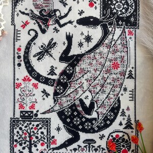 May include: A cross-stitch embroidery on white fabric featuring a large black dragon with patterned wings, a small fairy, dragonflies, and trees. The design uses black, white, and red thread to depict a forest scene with stars and mushrooms.
