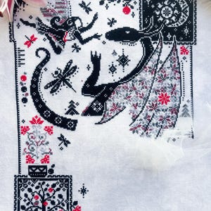 May include: A white fabric piece featuring a black, red, and gray cross-stitch pattern. The design includes a winged creature, a fairy, dragonflies, a tree, and a moon. The intricate embroidery displays geometric borders and floral motifs.