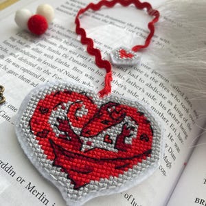 Peut inclure: Marque-page fait main en forme de c&oelig;ur avec un motif de dragon rouge, brod&eacute; au point de croix sur fond gris. Le marque-page a un ruban rouge et est pos&eacute; sur un livre ouvert.