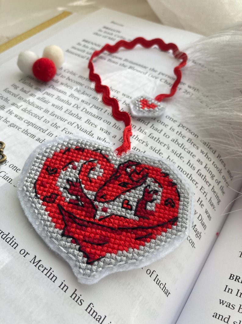 Peut inclure: Marque-page fait main en forme de c&oelig;ur avec un motif de dragon rouge, brod&eacute; au point de croix sur fond gris. Le marque-page a un ruban rouge et est pos&eacute; sur un livre ouvert.