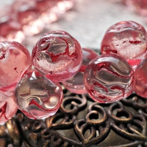 May include: A strand of pink glass beads with red swirls inside. The beads are round and have a translucent appearance.